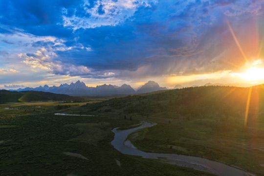 Aerial view of Jackson Hole ranchland at sunset, a winding river through green meadows beneath the Teton range and a break of golden storm light.