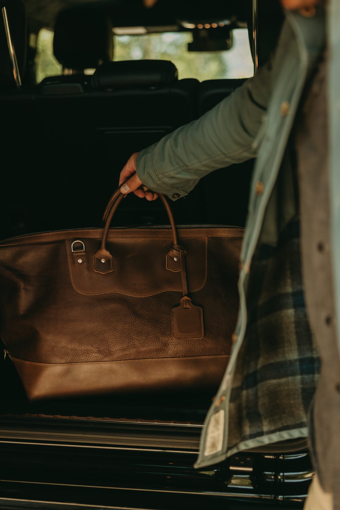 Brown leather travel bag being lifted from the rear of a vehicle.