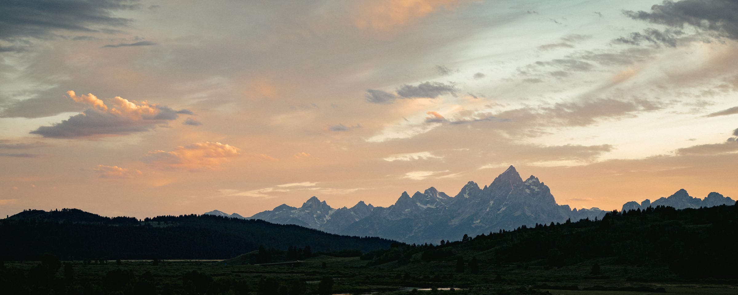 The Teton Range at dusk, the Grand Teton rising above a dark valley as a river bends through the foreground under a soft pink and gold sky.