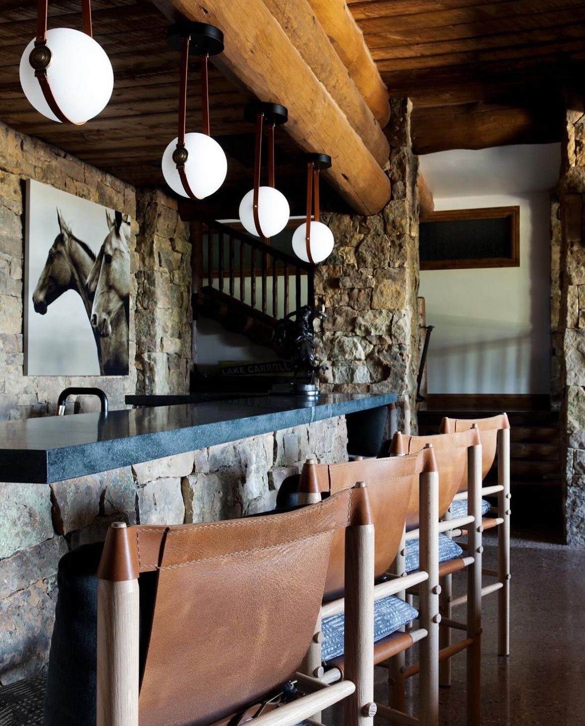 A rustic-modern ranch lounge with stone walls, black-and-white equestrian photography, leather barstools, and a honed stone bar beneath globe pendant lights